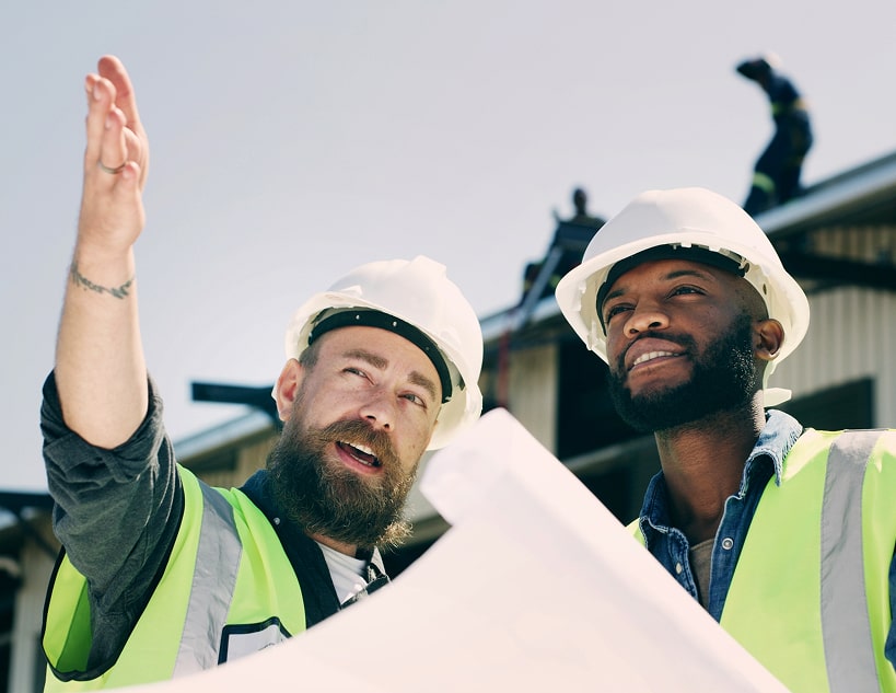Two construction workers in hard hats and safety vests examine a blueprint.