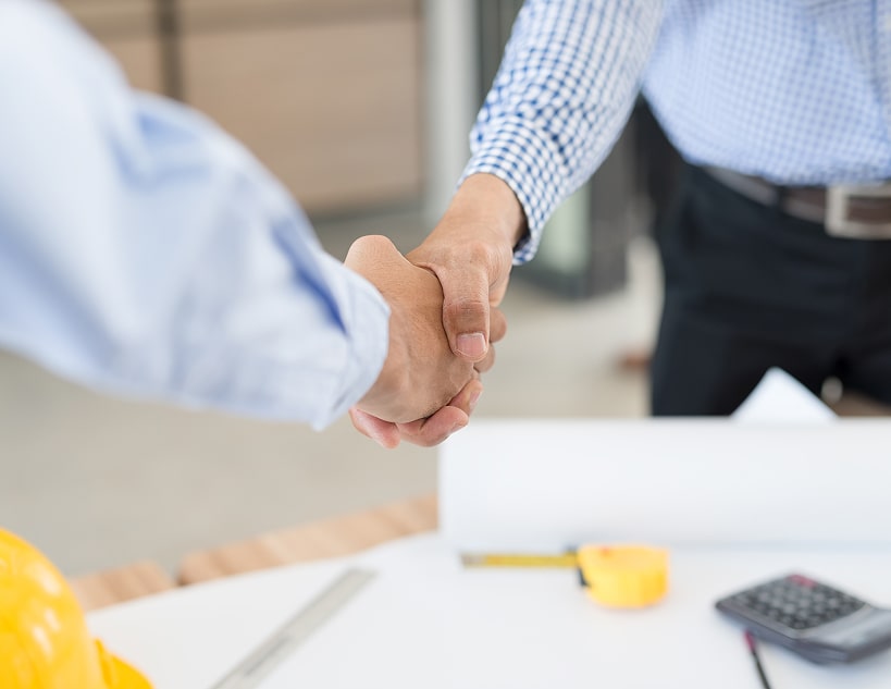 Two people shaking hands over a table with construction plans and tools.