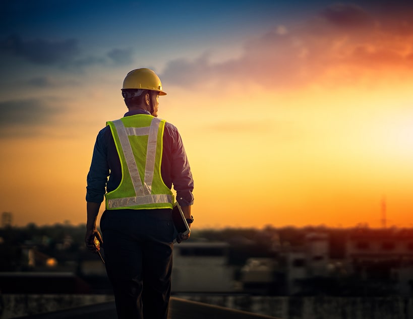 Construction worker in hard hat and safety vest stands on rooftop, looking out over city at sunset.