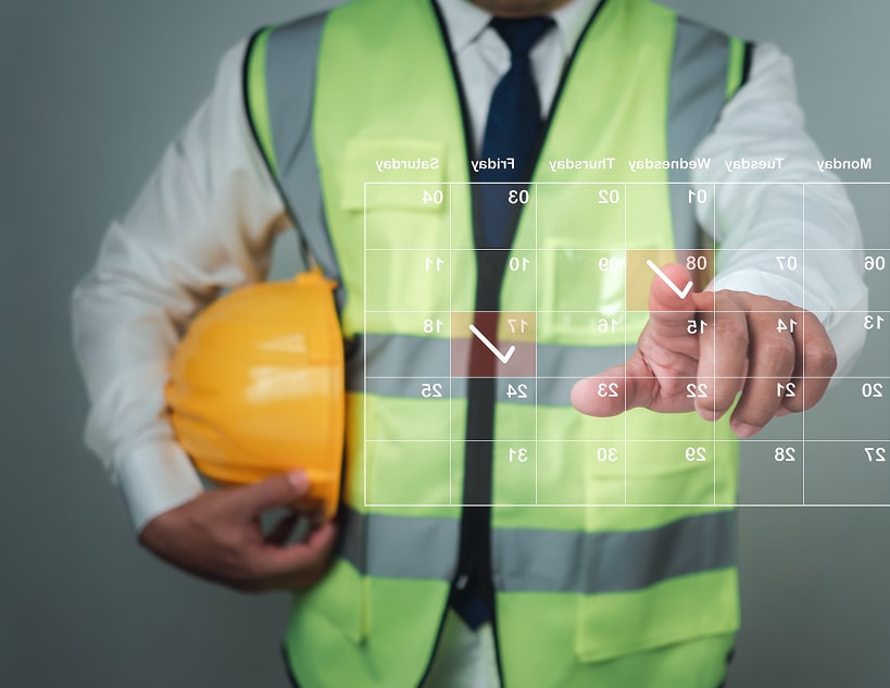 A man in a hard hat and safety vest, with a calendar superimposed over him. The man is wearing a white shirt, dark blue tie, and a neon yellow safety vest with reflective gray stripes. He holds an orange hard hat in his right hand and points to the calendar with his left index finger.