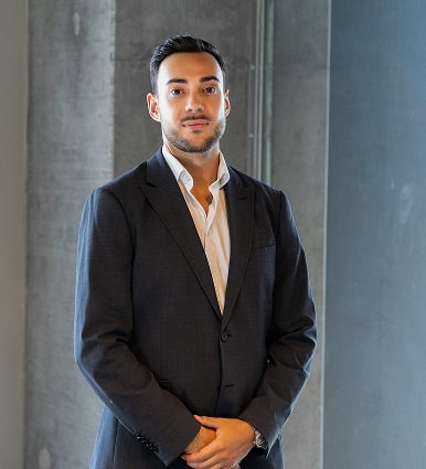 Man in a black suit jacket and white shirt standing in front of a concrete wall.